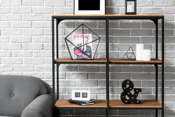 Shelf unit with stylish decor, magazine, alarm clock and candles near grey brick wall