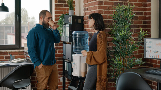Colleagues on break having discussion and using water cooler in business office. Man and woman talking while drinking cup of water as refreshment, using liquid dispenser for hydration
