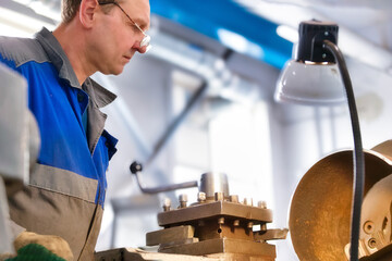 Elderly turner of 50-55 years old in overalls works at lathe in production shop. Processing of metal products. Real portrait of Caucasian worker. Production background.