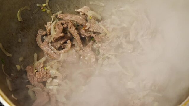 Slices of meat and onions are frying in the cauldron. The liquid in the wok is boiling and steam is rising. Caucasian cuisine