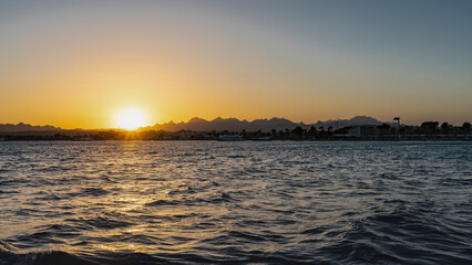 Fototapeta premium Sunset on the Red Sea. The rays of the setting sun illuminate a picturesque mountain range. The sky is colored orange. Waves on the water. Egypt. Safaga