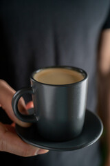 A man's hand holds a cup of hot espresso coffee against the background of a black T-shirt.