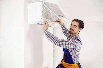 Portrait of happy young repairman fixing AC at work. Smiling handsome technician in workwear uses...