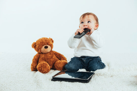 Studio photo with the white background of a baby's face with a mobile in his mouth next to a teddy bear and a tablet