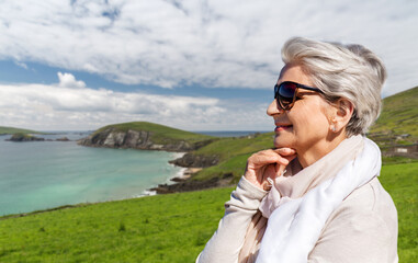 travel, tourism and vacation concept - portrait of happy senior woman in sunglasses and scarf over atlantic ocean coast in ireland background - obrazy, fototapety, plakaty
