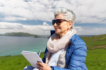 travel, tourism and vacation concept - happy senior woman writing to notebook over atlantic ocean coast in ireland background