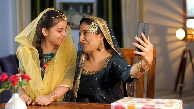 A young Muslim mother and her cute daughter clicking a selfie together on a mobile - modern lifestyle. A middle-aged pretty lady and a little girl-child wearing an Indian festive dress - Eid celebr...