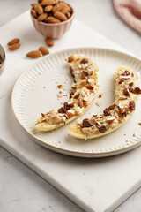 Two banana halves ready with peanut butter topping, raisins, almonds and chia seeds on white plate on marble board