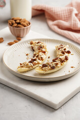 Two banana halves ready with peanut butter topping, raisins, almonds and chia seeds on white plate on marble board