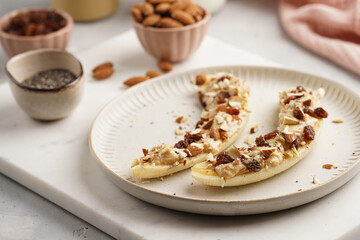 Two banana halves ready with peanut butter topping, raisins, almonds and chia seeds on white plate on marble board