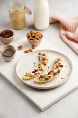 Two banana halves ready with peanut butter topping, raisins, almonds and chia seeds on white plate on marble board