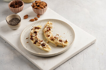 Two banana halves ready with peanut butter topping, raisins, almonds and chia seeds on white plate...