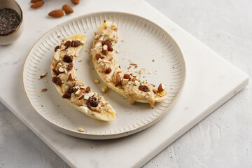 Two banana halves ready with peanut butter topping, raisins, almonds and chia seeds on white plate...