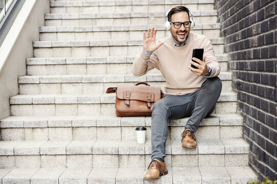 A happy urban man sits on the stairs outside, having video call on the phone.