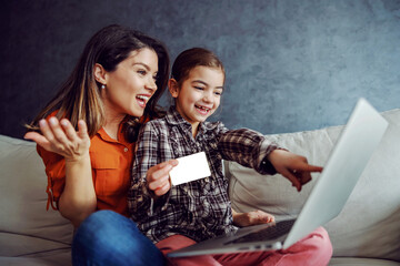 Fototapeta Smiling mother and daughter sitting on sofa and using laptop for online shopping.  obraz