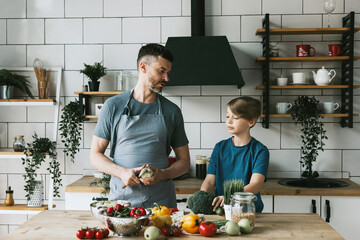 Fototapeta na wymiar family dad young man and son teenage boy cook vegetable salad in kitchen and spend quality time together, father and son talking and cooking vegetarian food and doing chores, 8 march and mothers day