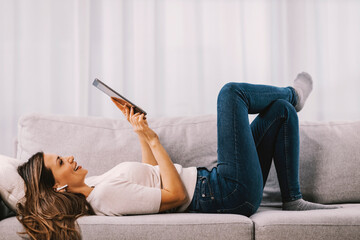A woman lies down on the sofa and scrolls on the tablet.