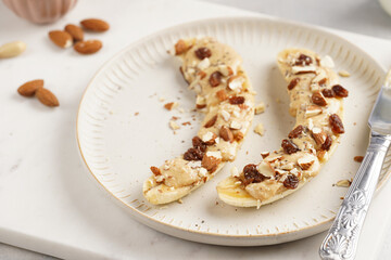 Two banana halves ready with peanut butter topping, raisins, almonds and chia seeds on white plate on marble board