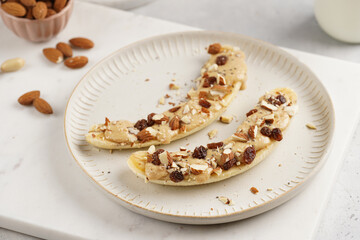 Two banana halves ready with peanut butter topping, raisins, almonds and chia seeds on white plate on marble board
