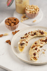 Two banana halves ready with peanut butter topping, raisins, almonds and chia seeds on white plate on marble board