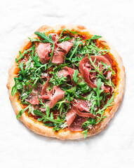 Homemade pizza with tomato sauce, prosciutto and arugula on a light background, top view