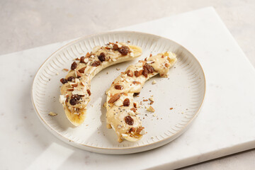 Two banana halves ready with peanut butter topping, raisins, almonds and chia seeds on white plate on marble board, direct creative sunlight shadow