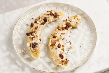 Two banana halves ready with peanut butter topping, raisins, almonds and chia seeds on white plate on marble board, direct creative sunlight shadow
