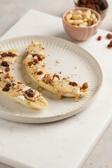 Two banana halves ready with peanut butter topping, raisins, almonds and chia seeds on white plate on marble board, direct creative sunlight shadow