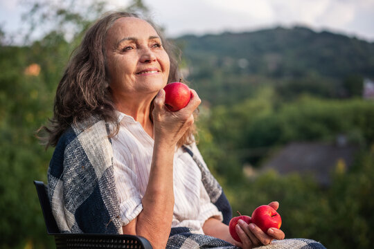 Cheerful beautiful happy mature senior woman holding red apples sitting in her countru house garden