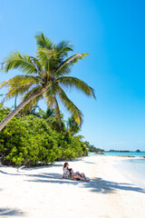 Woman and child lying in the shade of a palm tree on a white sand beach