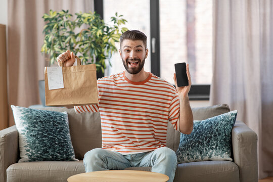technology, delivery and people concept - smiling man with smartphone and takeaway food in paper bag at home