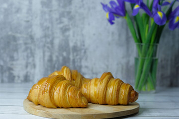 Sweet baked croissants on wooden cutting board on white wooden table with bouquet flowers of Irises and concrete wall background. 