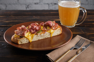 Crispy ciabatta bruschetta with bacon and melted cheese on a plate with cutlery and a mug of light beer on a wooden table. Horizontal orientation, copy space, no people.