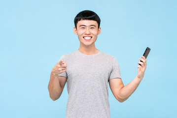 Positive Asian male in casual wear holding cellphone while pointing at camera against light blue isolated background in studio