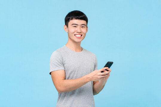 Positive Asian man in casual wear texting on cellphone  with smile in light blue isolated background in studio