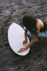 Young woman surfer, preparing a surfboard on the ocean, waxing. Woman with surfboard on the ocean, active lifestyle, water sports.