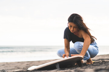 Young woman surfer, preparing a surfboard on the ocean, waxing. Woman with surfboard on the ocean, active lifestyle, water sports.