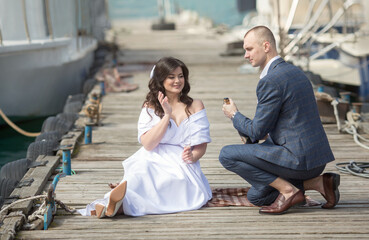 a young man in a suit and a woman in a white dress are sitting on a pier by the sea and drinking champagne, they are having a wedding.