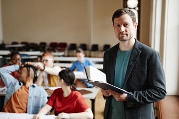 Waist up portrait of male college professor looking at camera in classroom hall and holding book,...