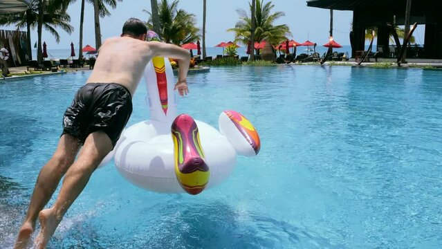 Happy funny man successfully jump onto floating inflatable unicorn in swimming pool in tropical hotel to ride and have fun, show yes rock sign symbol gesture by hand. Summer vacation, holiday concept.