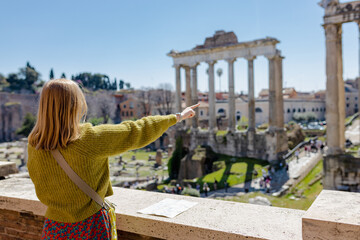 A happy blond woman tourist is standing near the Roman Forum, old ruins at the center of Rome, Italy. Concept of traveling famous landmarks. Girl pointing finger, sunny day - obrazy, fototapety, plakaty