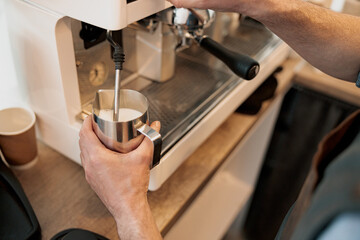 Barista regulating the supply of steam with tap on coffee machine, heating up milk in pitcher with...