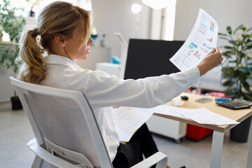 Female accountant examines documents with financial diagram and charts while sitting in a chair at the workplace