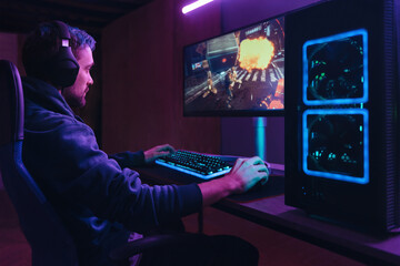 Young concentrated cyber sportsman playing in FPS video game on his personal computer in dark neon...