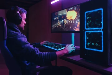Fotobehang Gaming Young concentrated cyber sportsman playing in FPS video game on his personal computer in dark neon room. Pro gamer participating in online esport competition. Cyber games championship event concept  © Arsenii