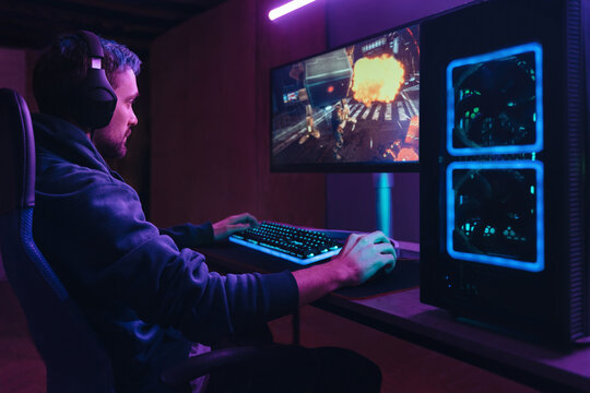 Young concentrated cyber sportsman playing in FPS video game on his personal computer in dark neon room. Pro gamer participating in online esport competition. Cyber games championship event concept