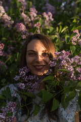 Portrait of attractive smiling caucasian woman surrounded by purple lilac flowers in the garden. Outdoor fashion photo.