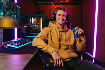 Portrait of smiling pro gamer wearing headset, looking at camera, holding energy caffeine dink, sitting on gaming chair near professional gaming setup. Cyber sportsman playing shooter computer game