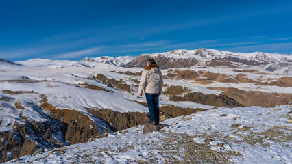 Naklejka premium A man in a down jacket stands on a high-altitude plateau at the edge of the gorge and looks into the distance. Back view. The red-brown slopes of the mountains are snow covered. Blue sky. Altai Mars 