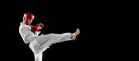 Male taekwondo fighter in white dobok, helmet and gloves training isolated over dark background....